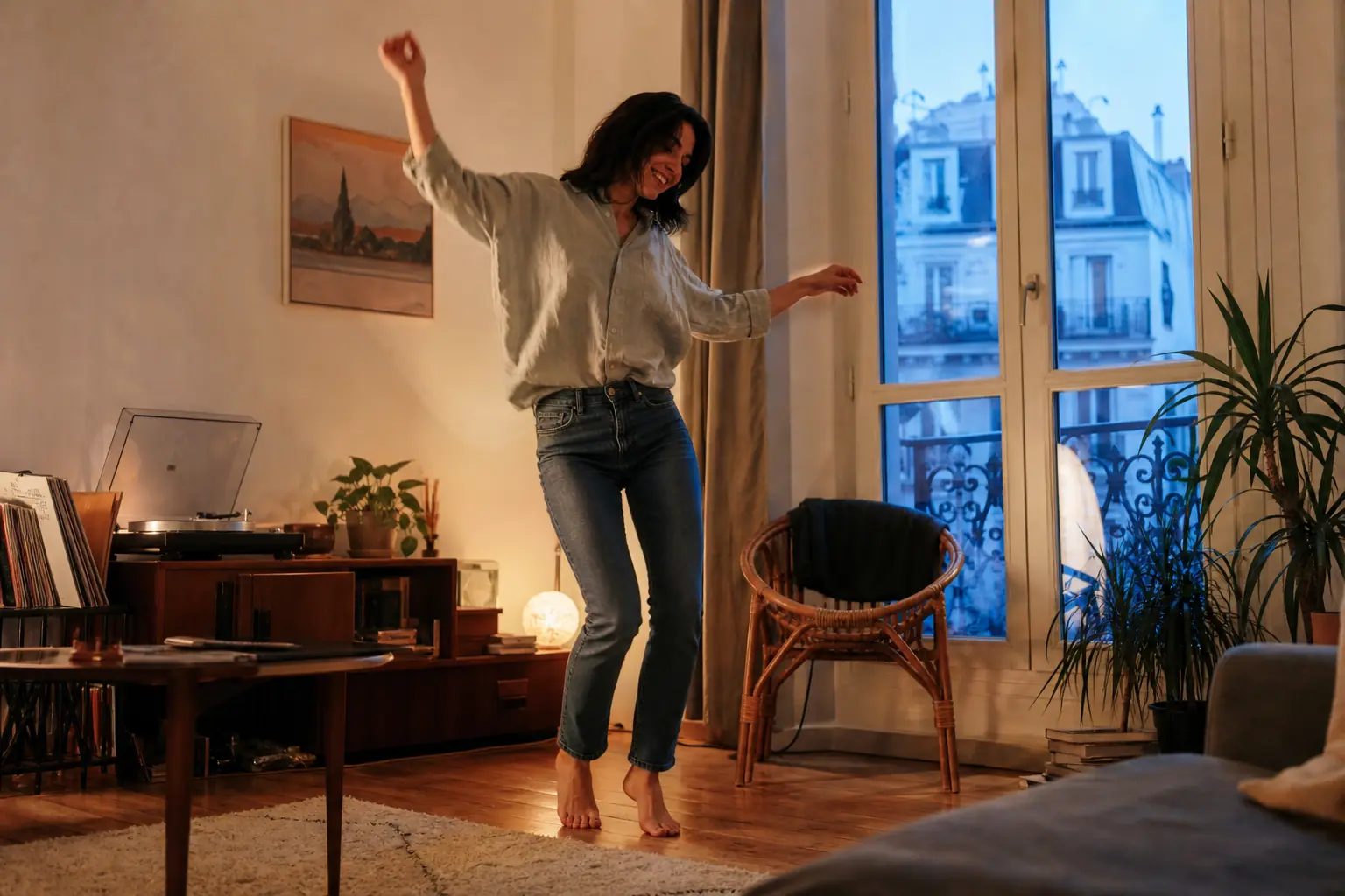 Une femme adulte danse pieds nus dans son salon, en jean et chemise, la lumière du soir entre par la fenêtre