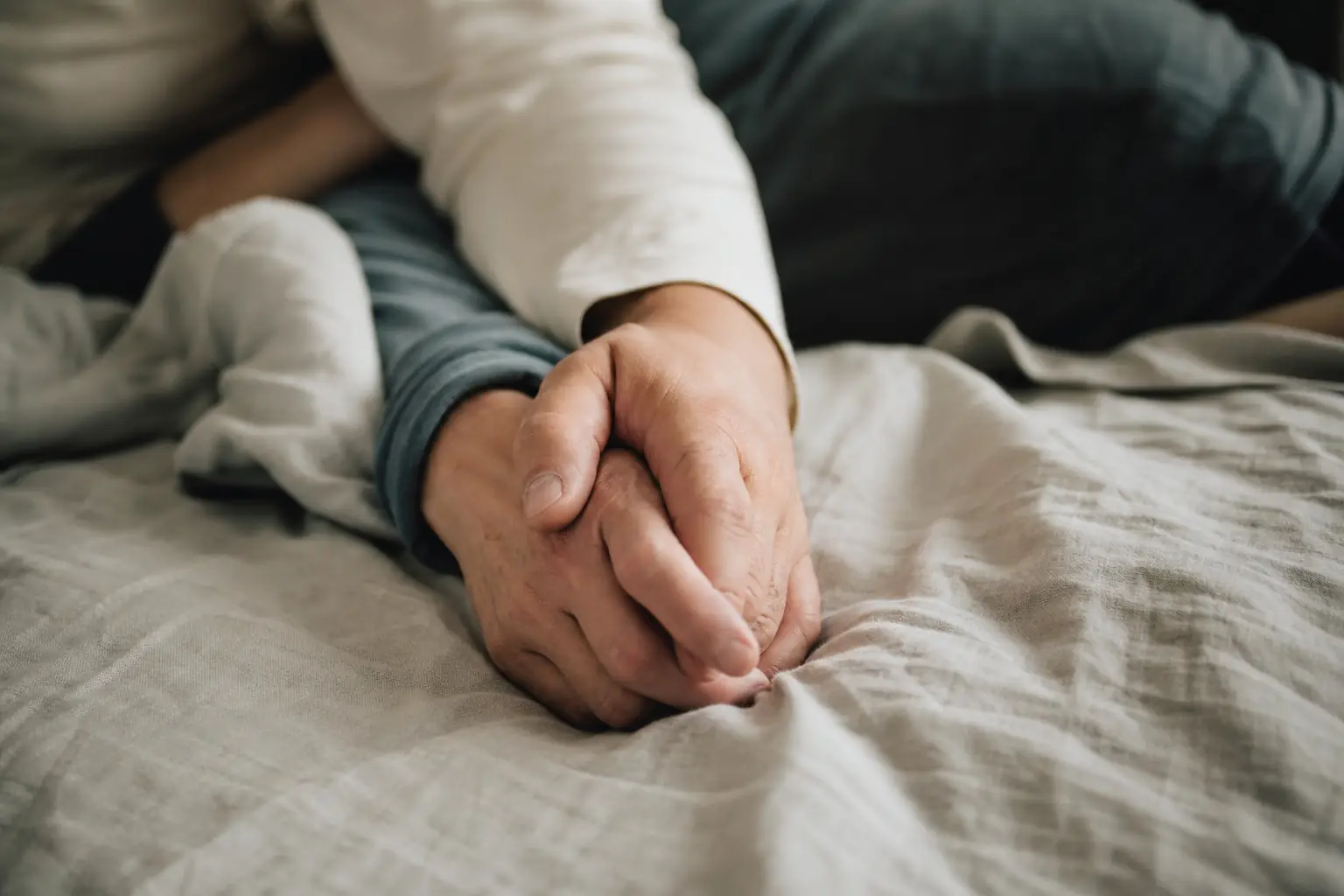 Deux mains adultes entrelacées sur un drap de lin froissé, lumière douce du matin