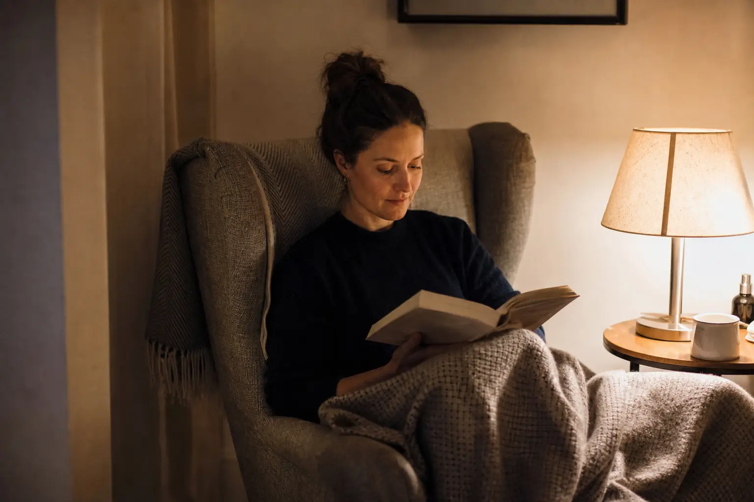 Une femme adulte lit un livre dans un fauteuil, une couverture sur les genoux