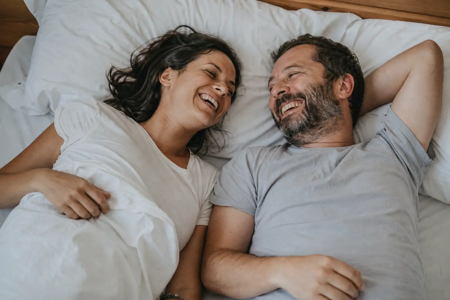 Un couple adulte allongé côte à côte, en tee-shirt, riant dans la lumière du matin