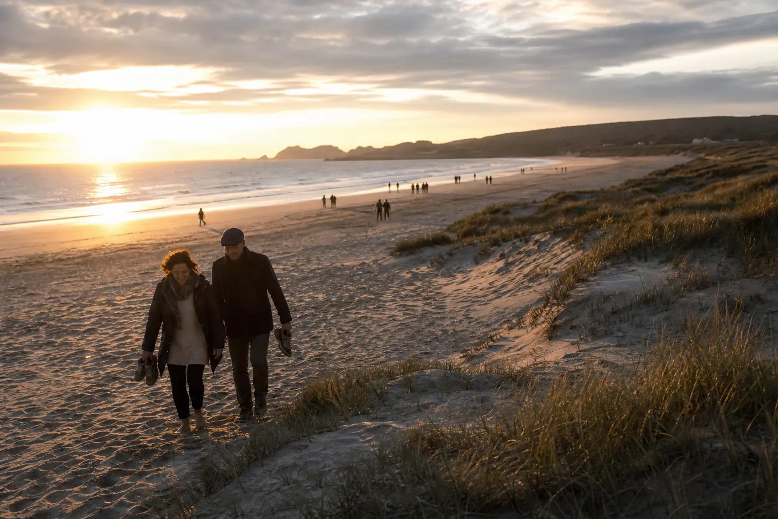 Une plage de sable au coucher du soleil, dunes et végétation, quelques promeneurs au loin