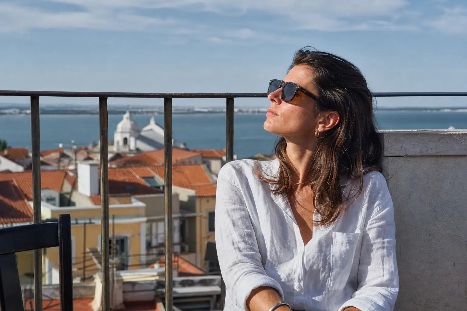 Une femme adulte porte des lunettes de soleil sombres sur une terrasse ensoleillée, le visage tourné vers la lumière